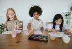 talented diverse little girls painting on papers with watercolors while sitting together at table