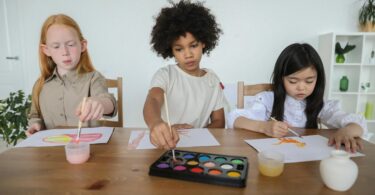 talented diverse little girls painting on papers with watercolors while sitting together at table